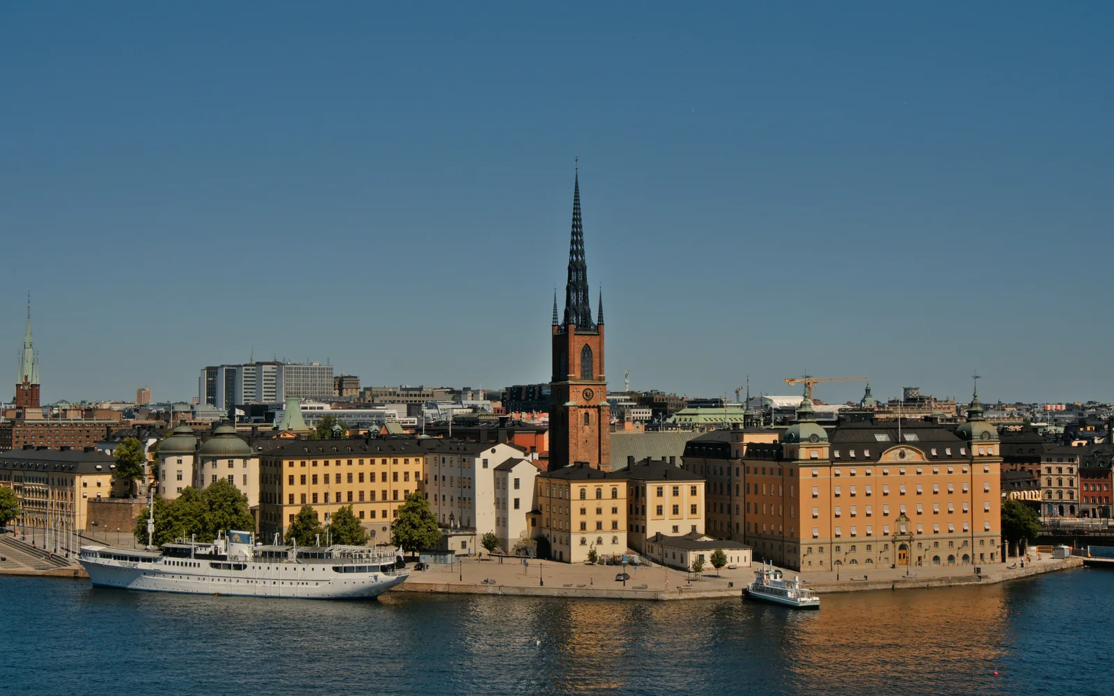 Riddarholmen i Stockholm sedd över vattnet, med Riddarholmskyrkans järnspira och gula fasader i kvällsljus